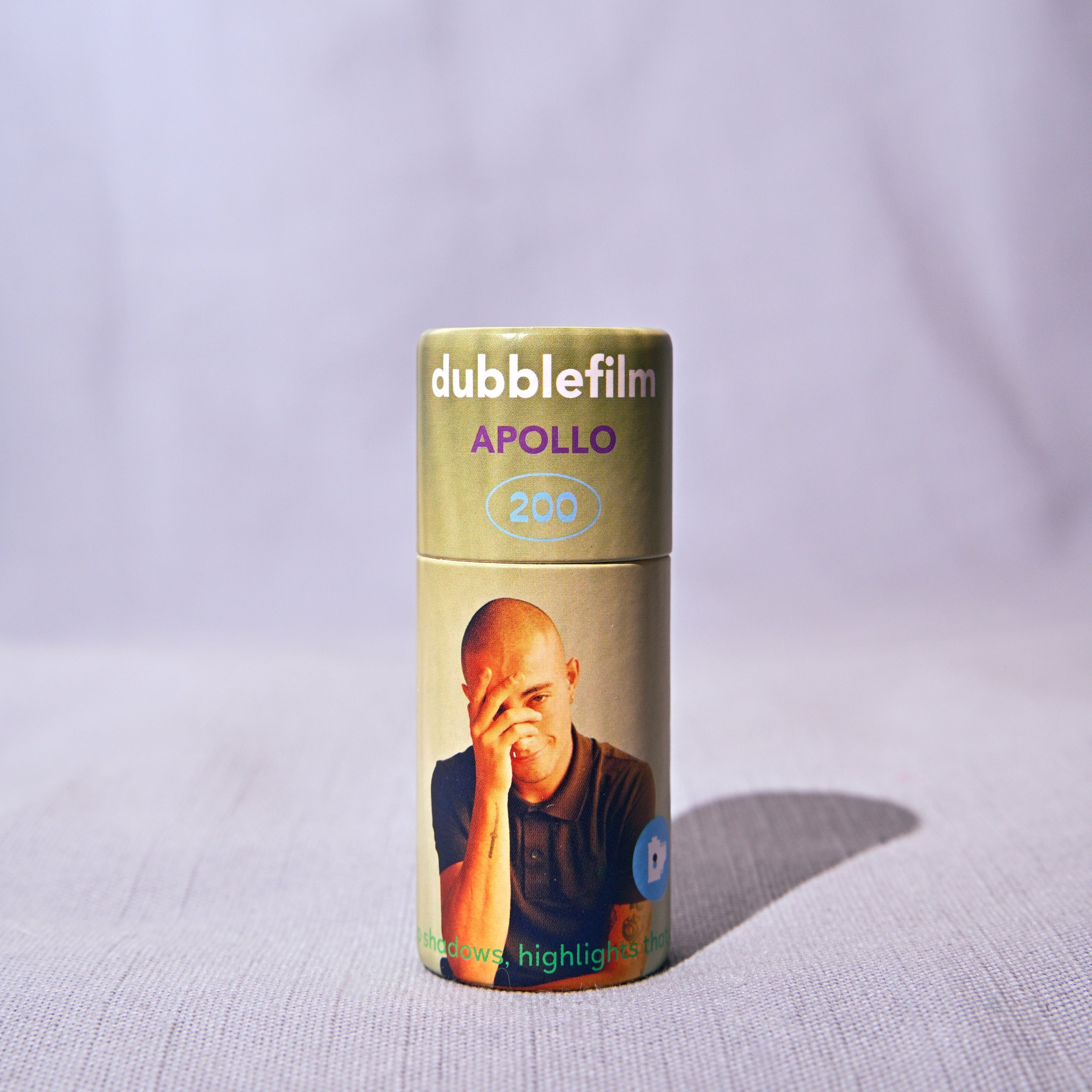 A single roll of doublefilm Apollo 200 color negative film stands upright on a light textured fabric surface against a draped light gray background.