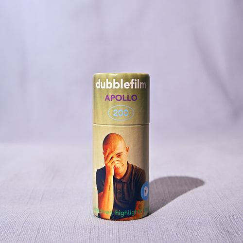 A single roll of doublefilm Apollo 200 color negative film stands upright on a light textured fabric surface against a draped light gray background.