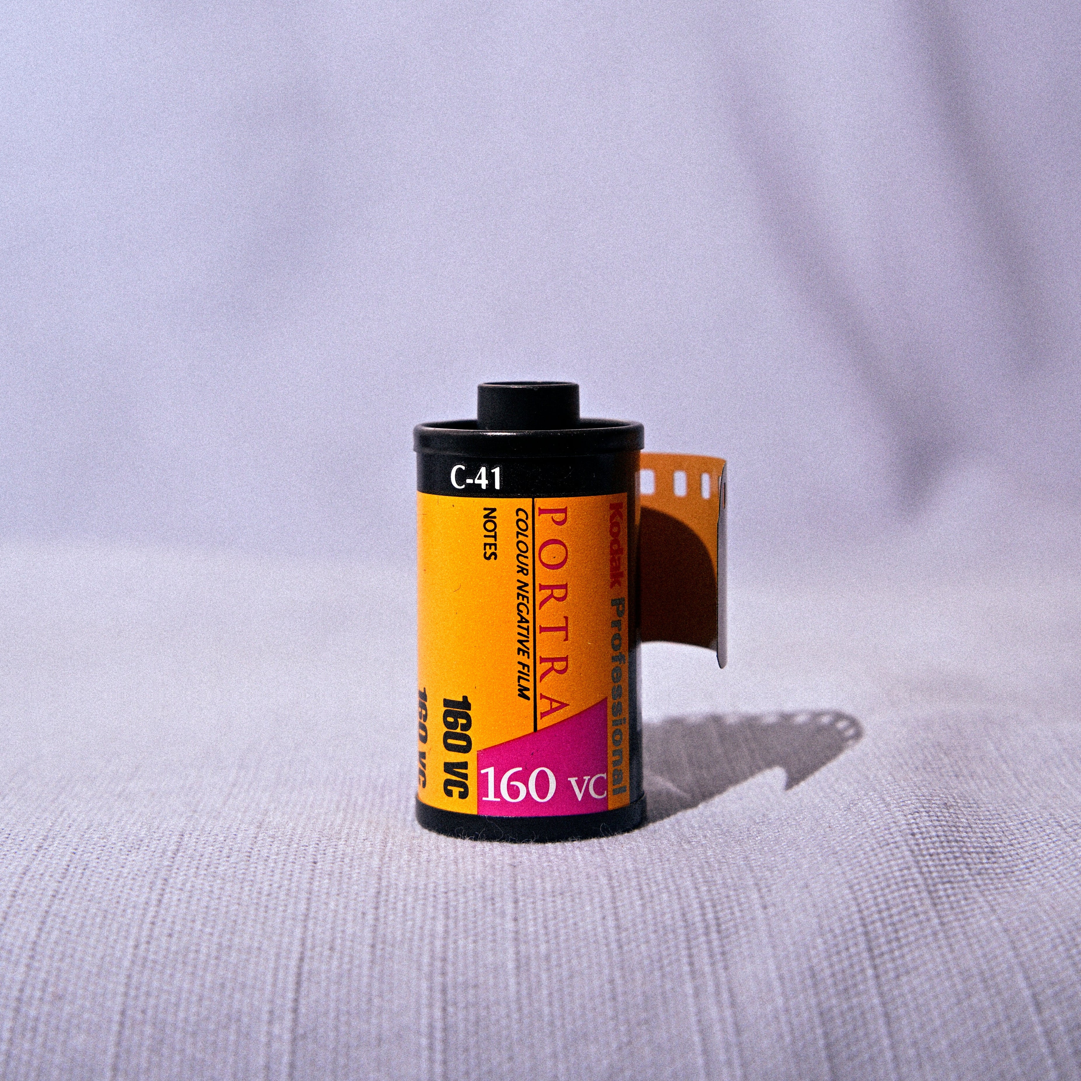 A single roll of Portra 160VC film with a yellow label and a strip of film partially unrolled, set against a soft, draped light purple fabric background.