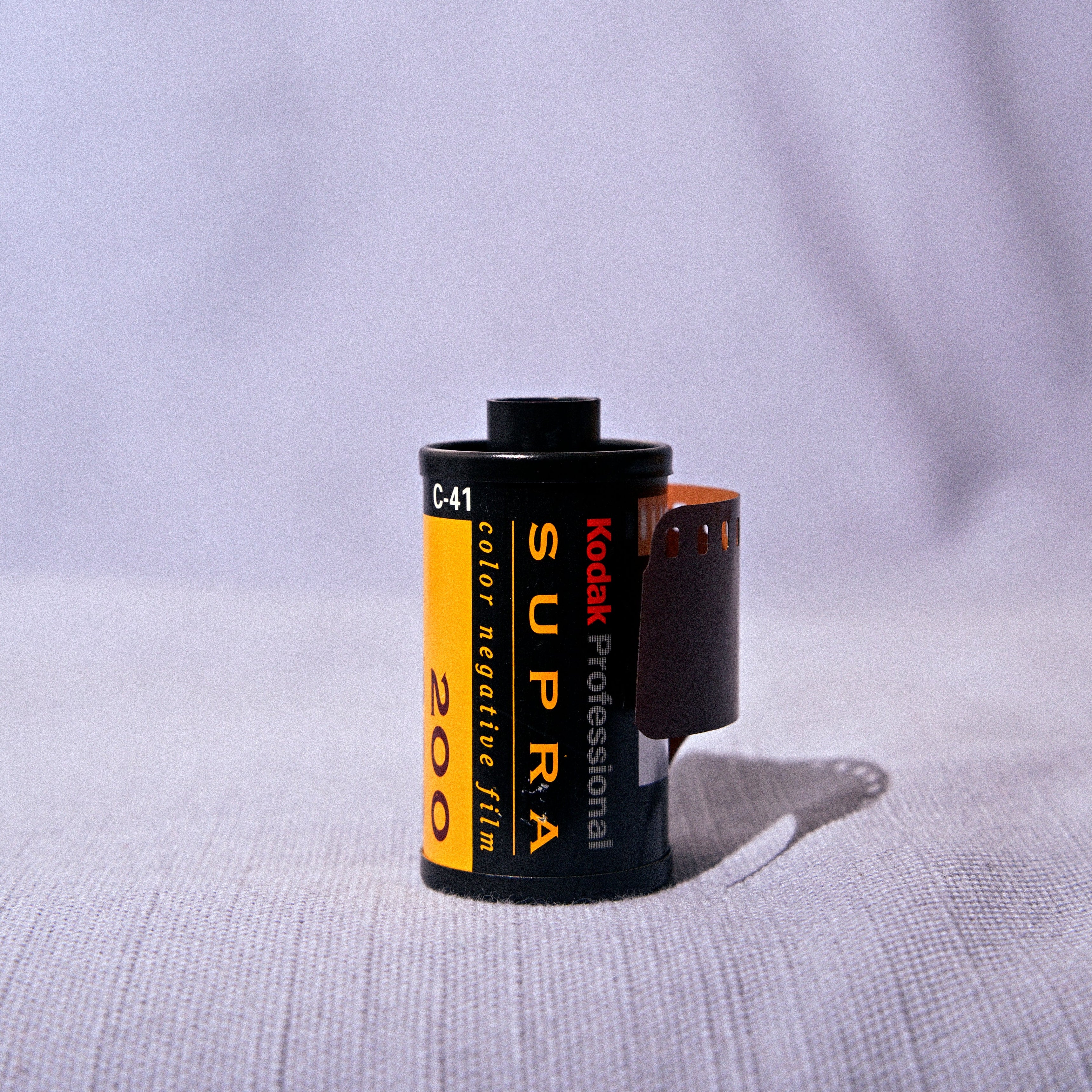 A single roll of Kodak Super 200 film partially unboxed, standing upright on a textured light gray fabric surface with a soft, draped background.