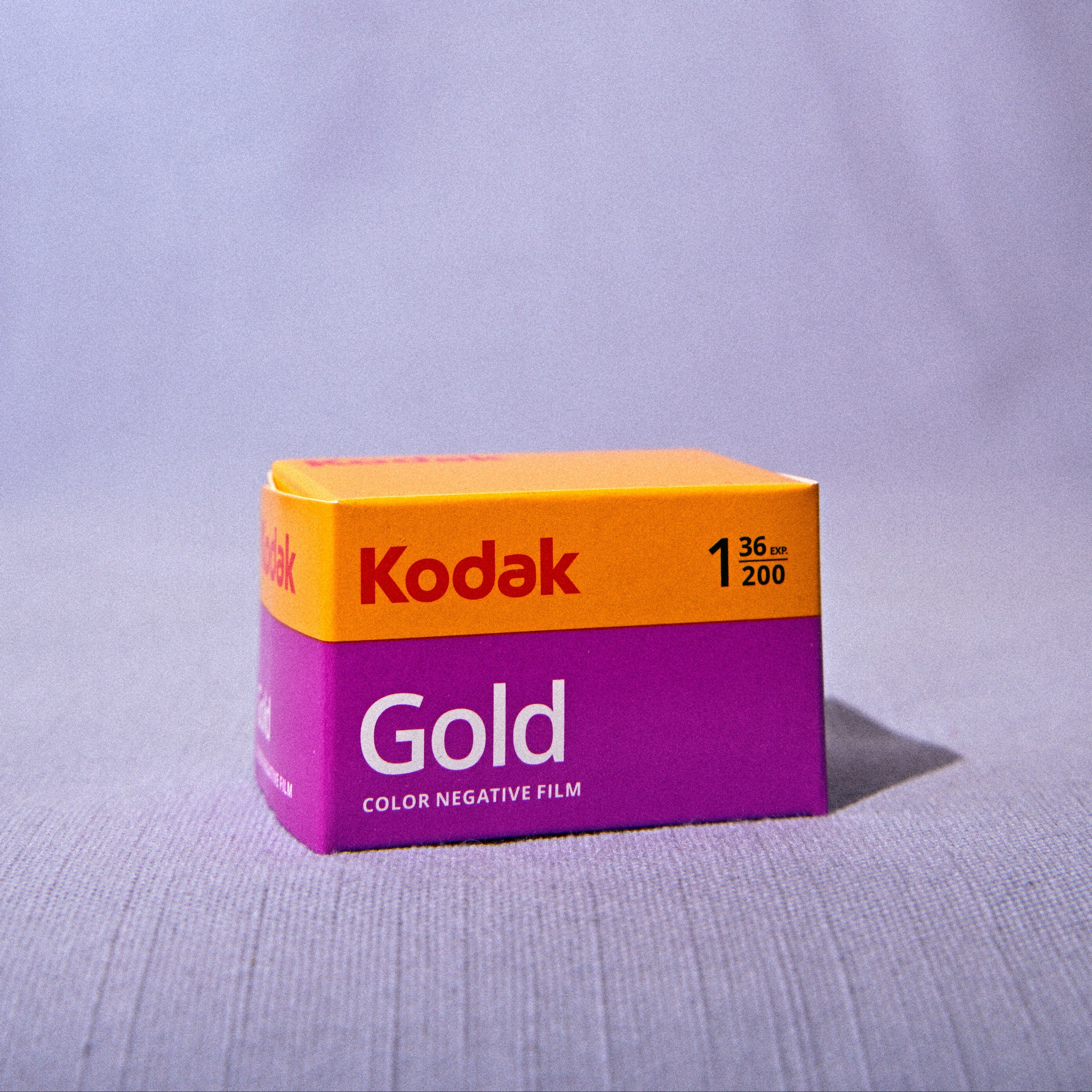 A box of Kodak Gold Color Negative Film, 35mm, standing upright on a textured light gray surface with a draped fabric backdrop.