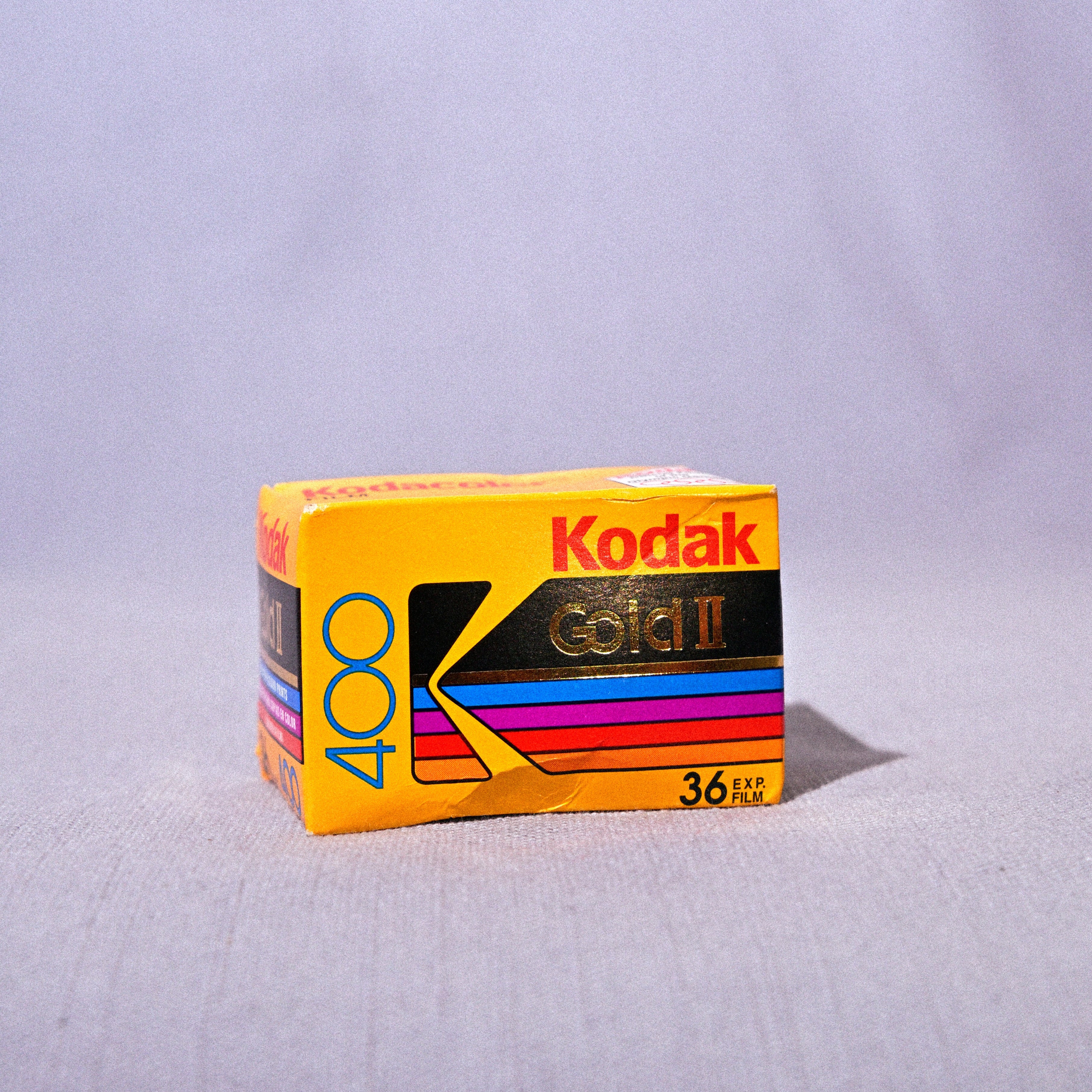 A vintage box of Kodak Gold 400 film, showing its iconic yellow and black packaging with colorful stripes, set against a light gray fabric backdrop.