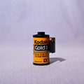 A single roll of Kodak Gold II film with a black and yellow label, standing upright on a light gray fabric surface with a matching soft gray background.