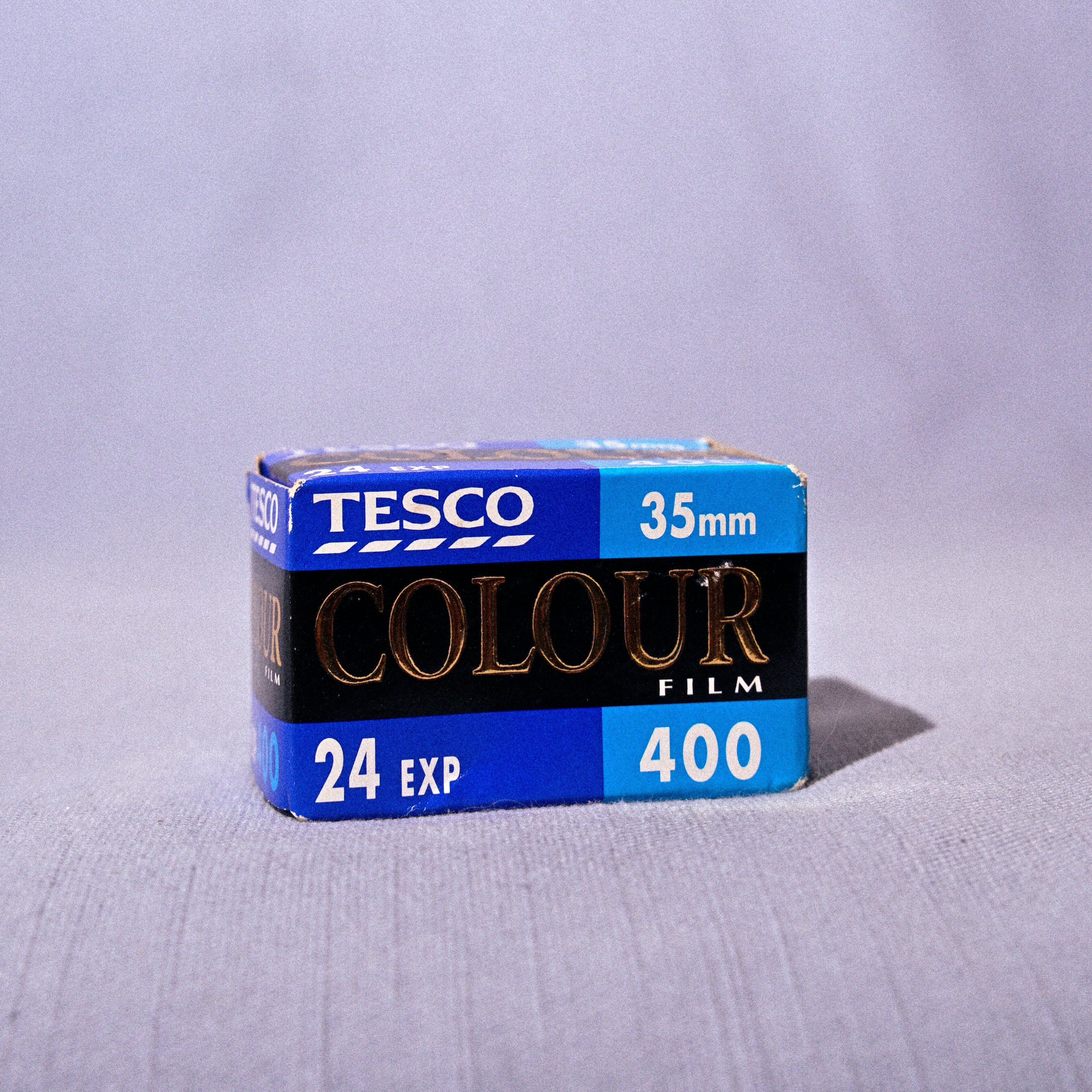 A box of Tesco Colour Film 24 Exp 400, 35mm, standing on a light gray fabric surface against a matching gray fabric backdrop.