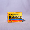 A vintage box of Kodacolor Gold 400 film, featuring its signature yellow and orange color scheme with black and gold lettering, set against a light gray fabric backdrop.