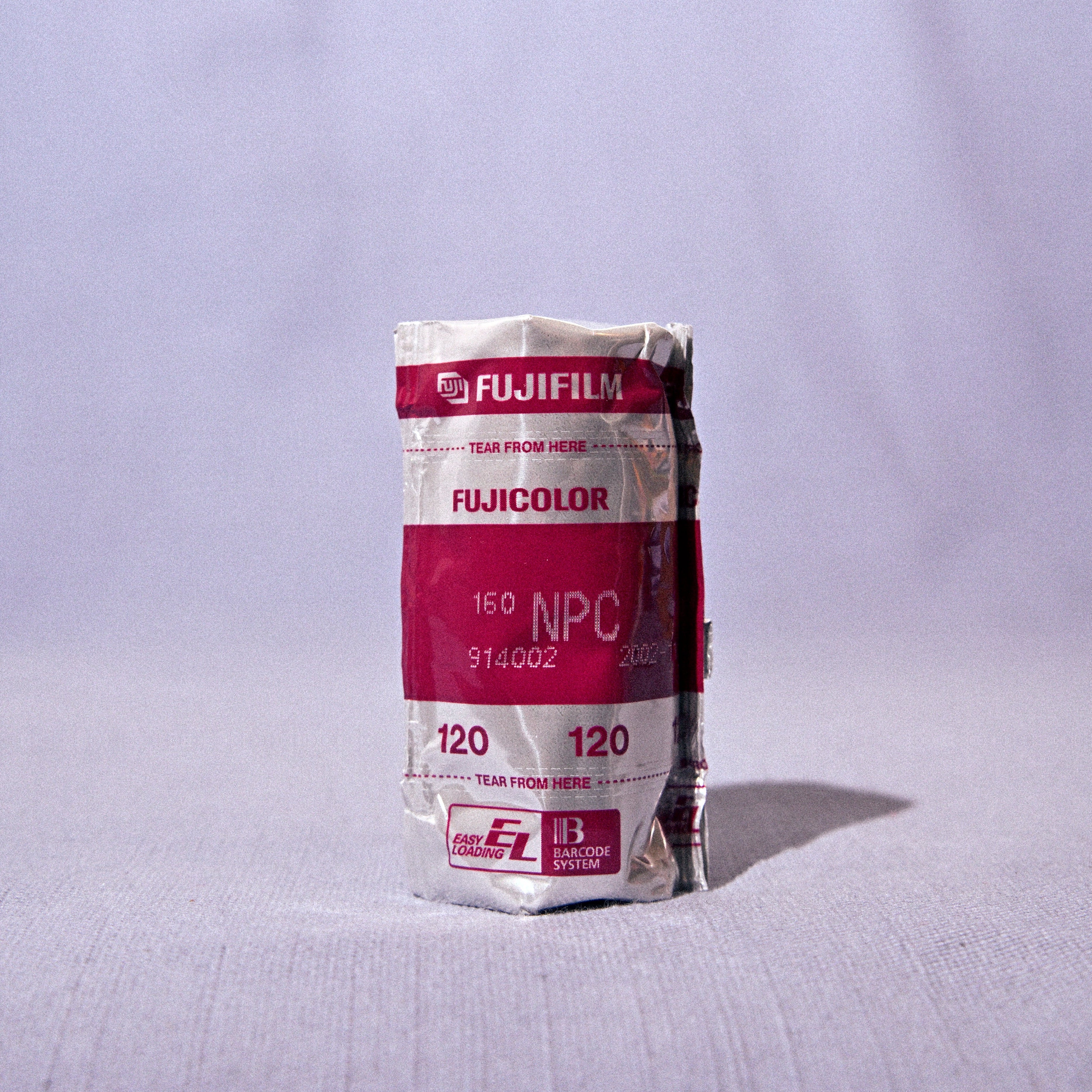 A roll of Fujifilm Color Neg 120 film, wrapped in its original paper packaging with a prominent magenta and white label, standing upright on a light gray textured surface against a matching light gray backdrop.