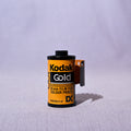A single roll of Kodak Gold DX 24mm film for color prints, standing upright on a light gray fabric surface with a soft shadow cast to the right.