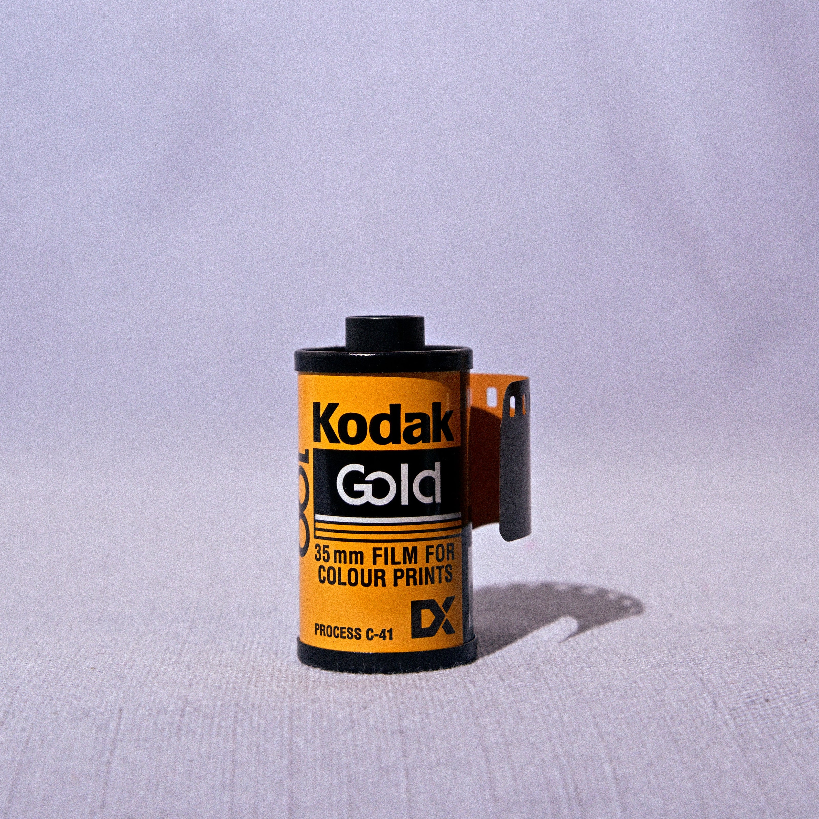 A single roll of Kodak Gold DX 24mm film for color prints, standing upright on a light gray fabric surface with a soft shadow cast to the right.
