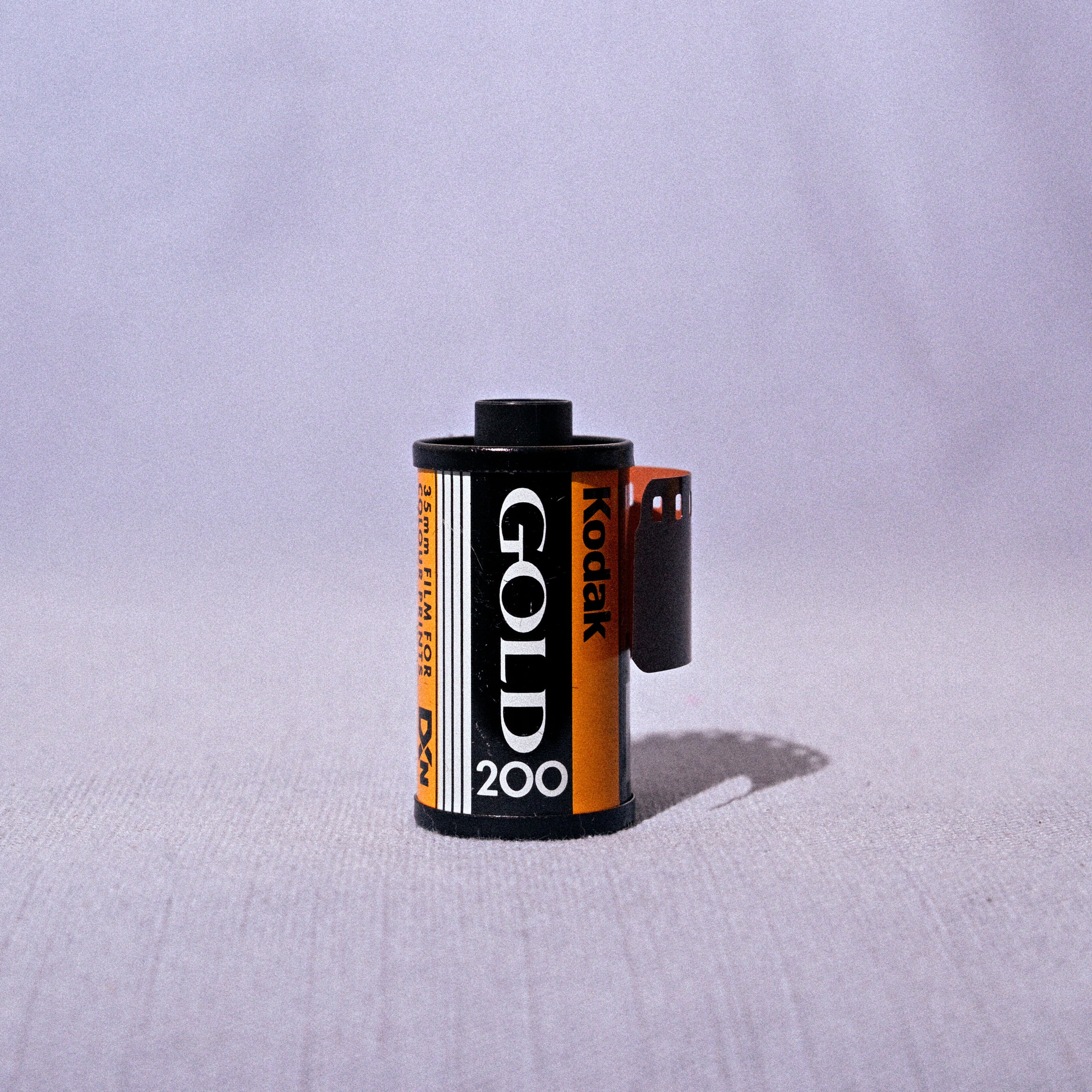 A roll of Kodak Gold 200 film partially unboxed, showing the black and orange label, set against a light gray fabric backdrop.