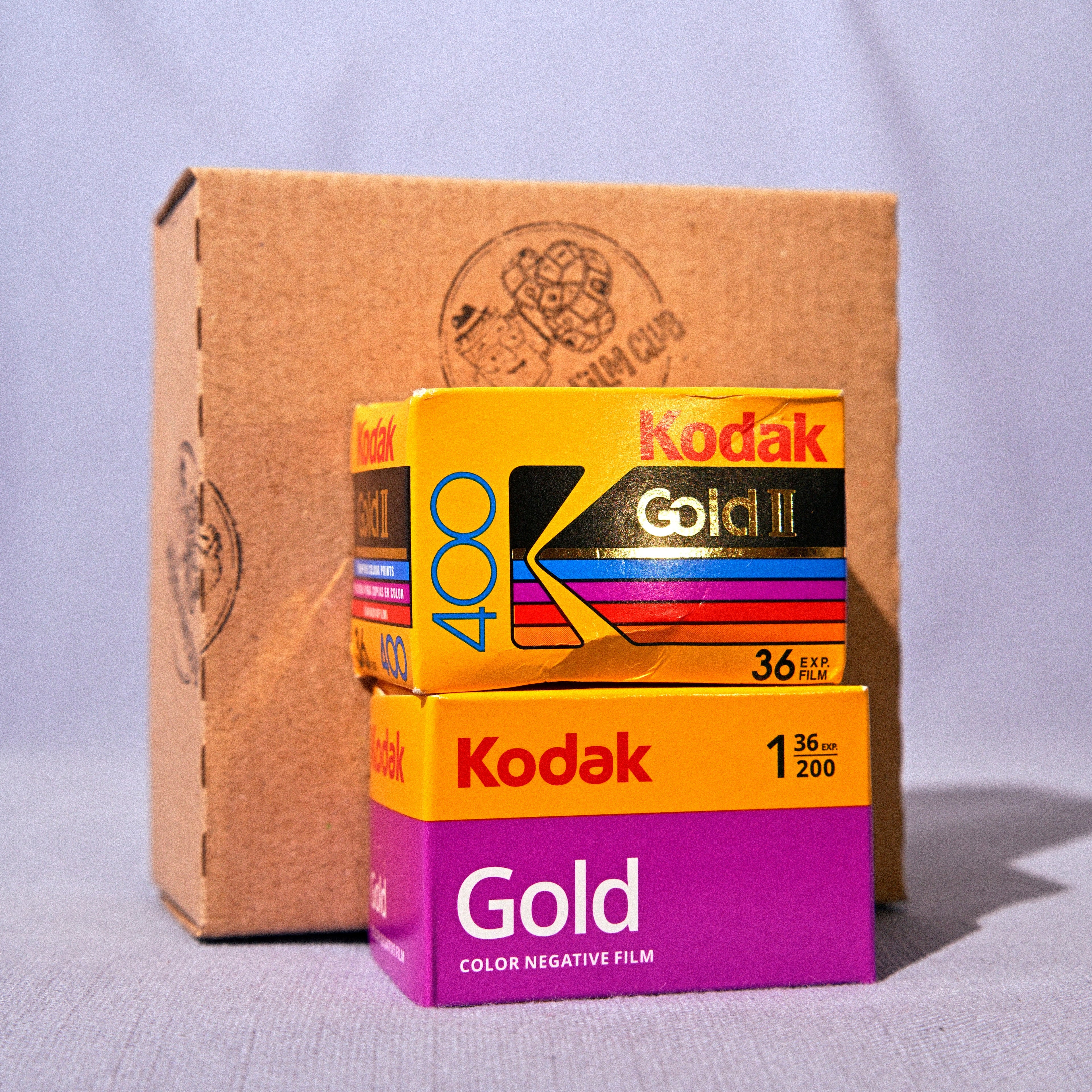 Two boxes of Kodak Gold Color Negative Film stacked in front of a large brown cardboard box, set against a light gray fabric backdrop.