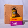 A vintage Kodak Gold 135 film canister displayed on top of its orange and purple box, with a larger brown cardboard box in the background, set against a light gray fabric backdrop.