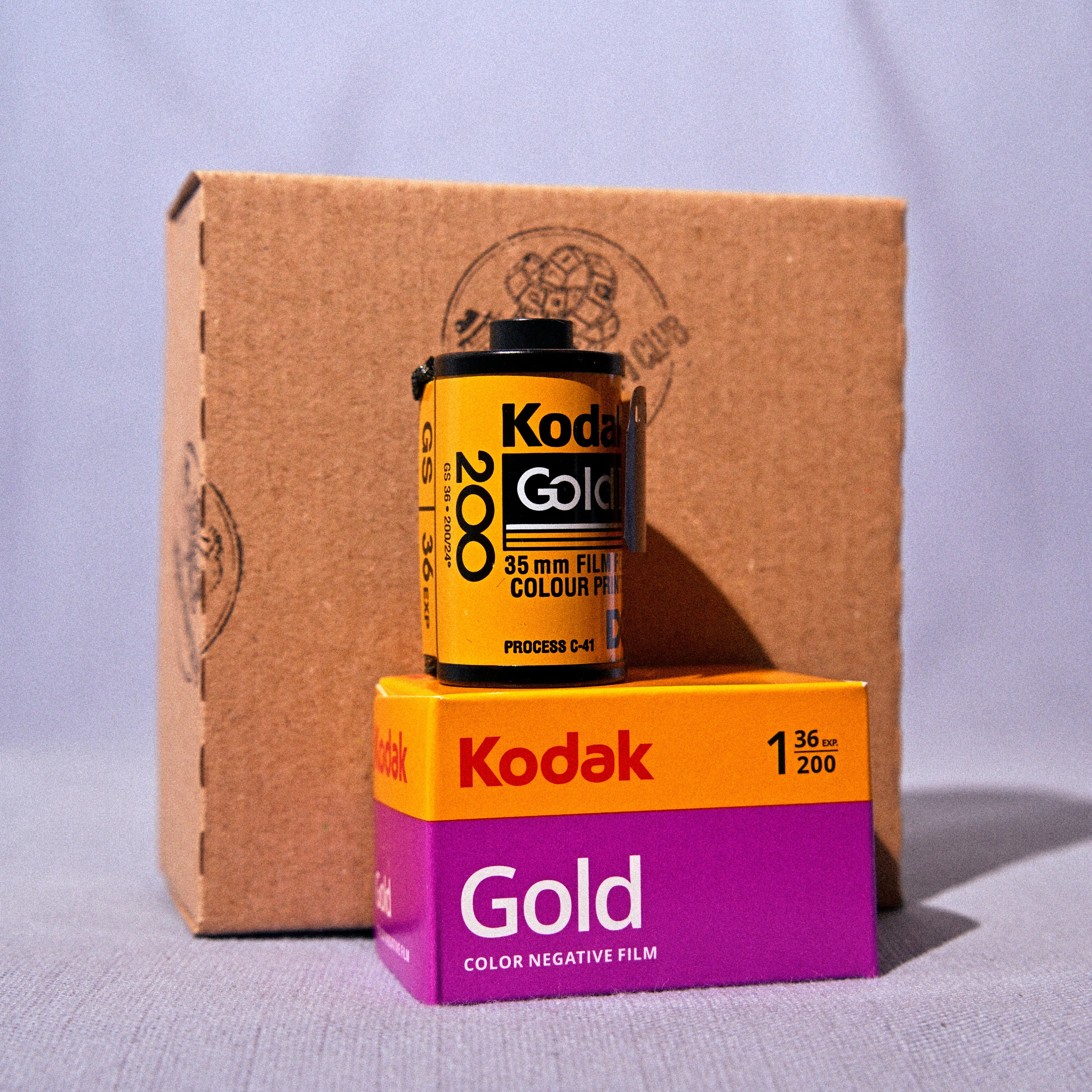 A vintage-style product display featuring a Kodak Gold 35mm film can resting on top of a Kodak Gold film box, with a large brown cardboard box in the background against a light gray fabric backdrop.