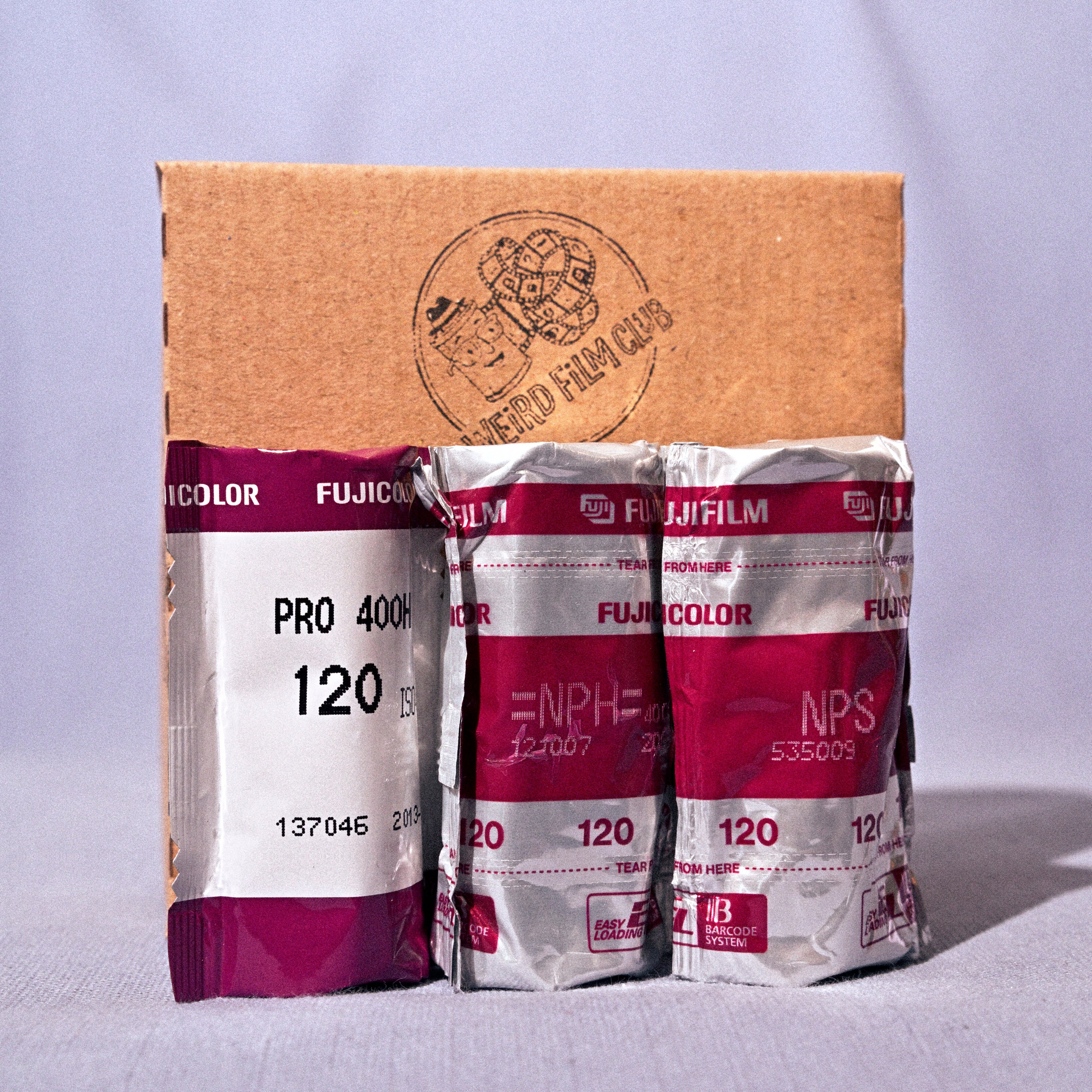 Three Fujicolor Pro 400 film packs, including one larger pack and two smaller packs, arranged in front of a brown cardboard box on a light gray fabric background.
