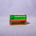 A vintage box of Fujicolor Super HR 100 film, featuring its signature green and brown packaging, set against a light gray fabric backdrop.