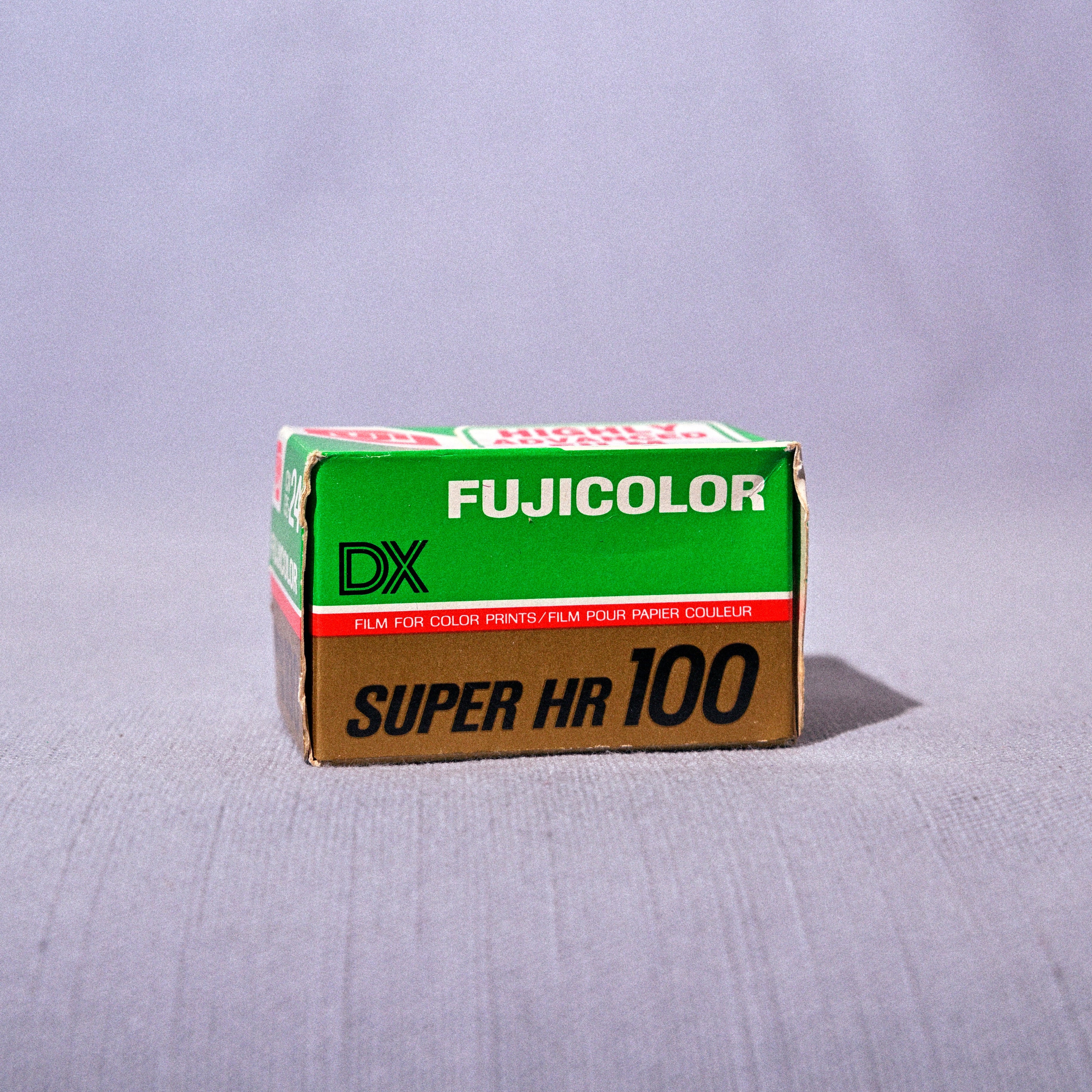 A vintage box of Fujicolor Super HR 100 film, featuring its signature green and brown packaging, set against a light gray fabric backdrop.