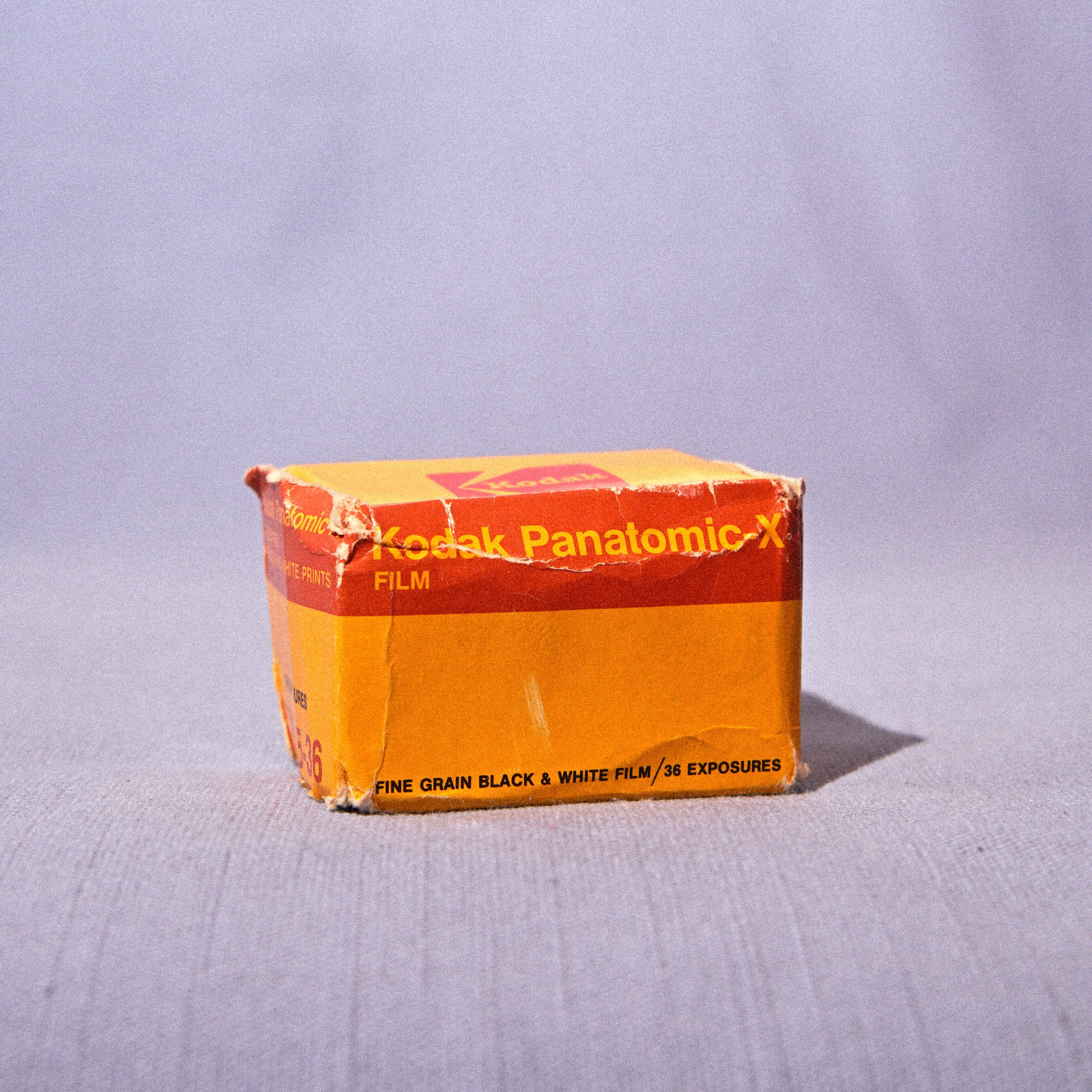 A vintage box of Kodak Panatomic-X film, showing its orange and yellow packaging, set against a plain light gray fabric backdrop.