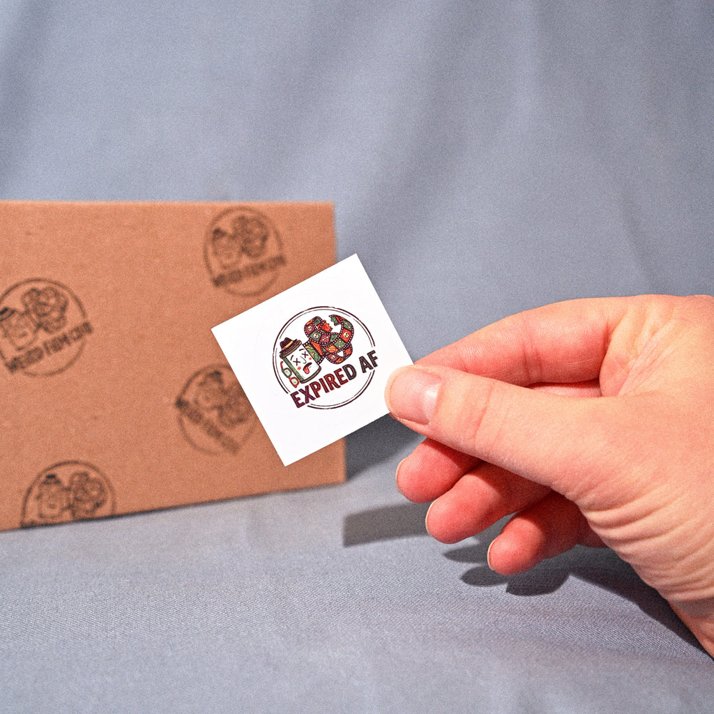 Hand holding a small white card with 'Expired AF' text next to a cardboard box on a gray background
