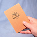 Hand holding a brown notebook with a logo and text on a light blue background