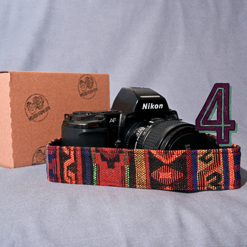 Camera with a colorful strap on a gray surface, next to a cardboard box and a number '4' sign.